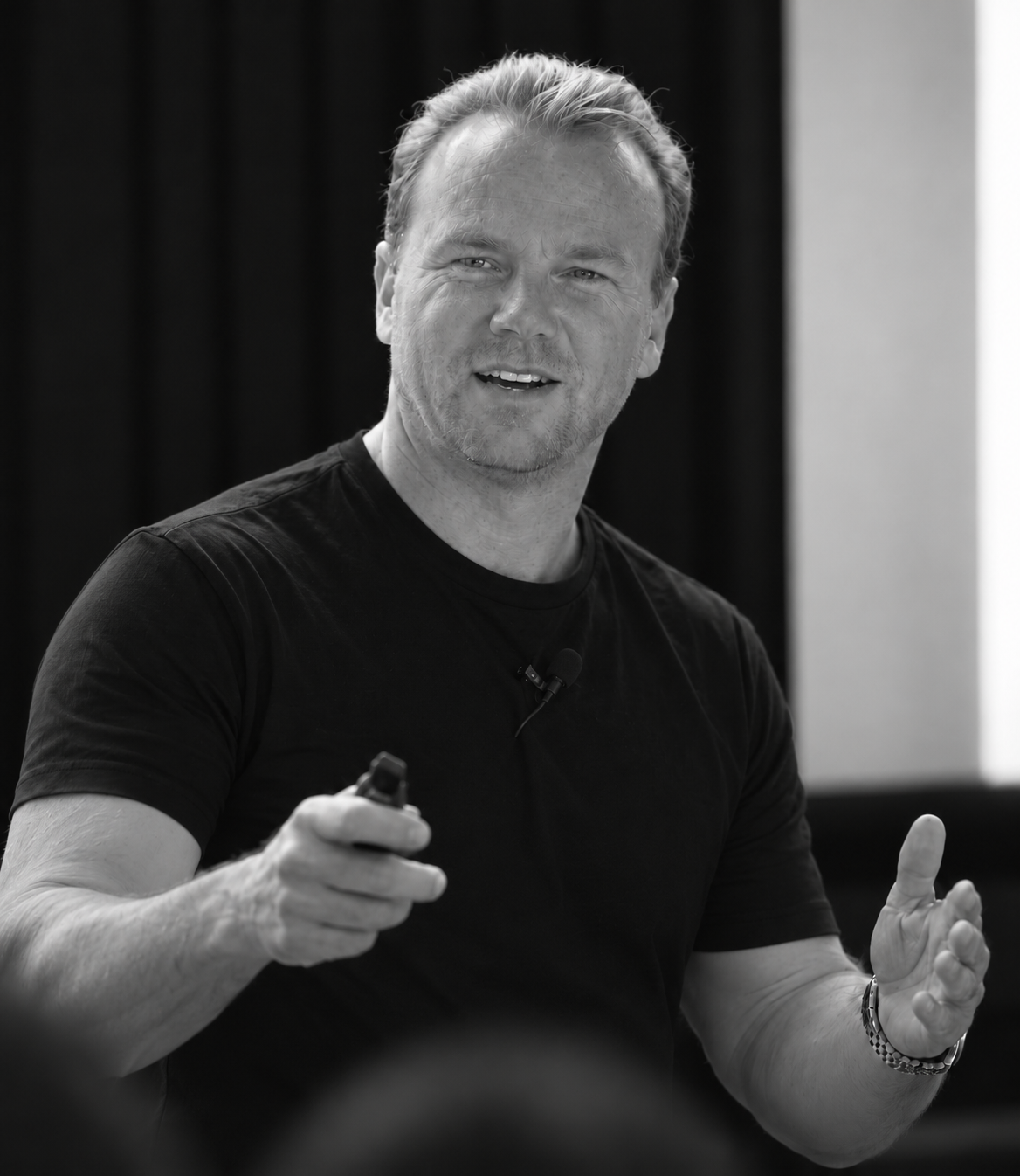 Richie Matthews presenting on stage in a black t-shirt, holding a slide clicker